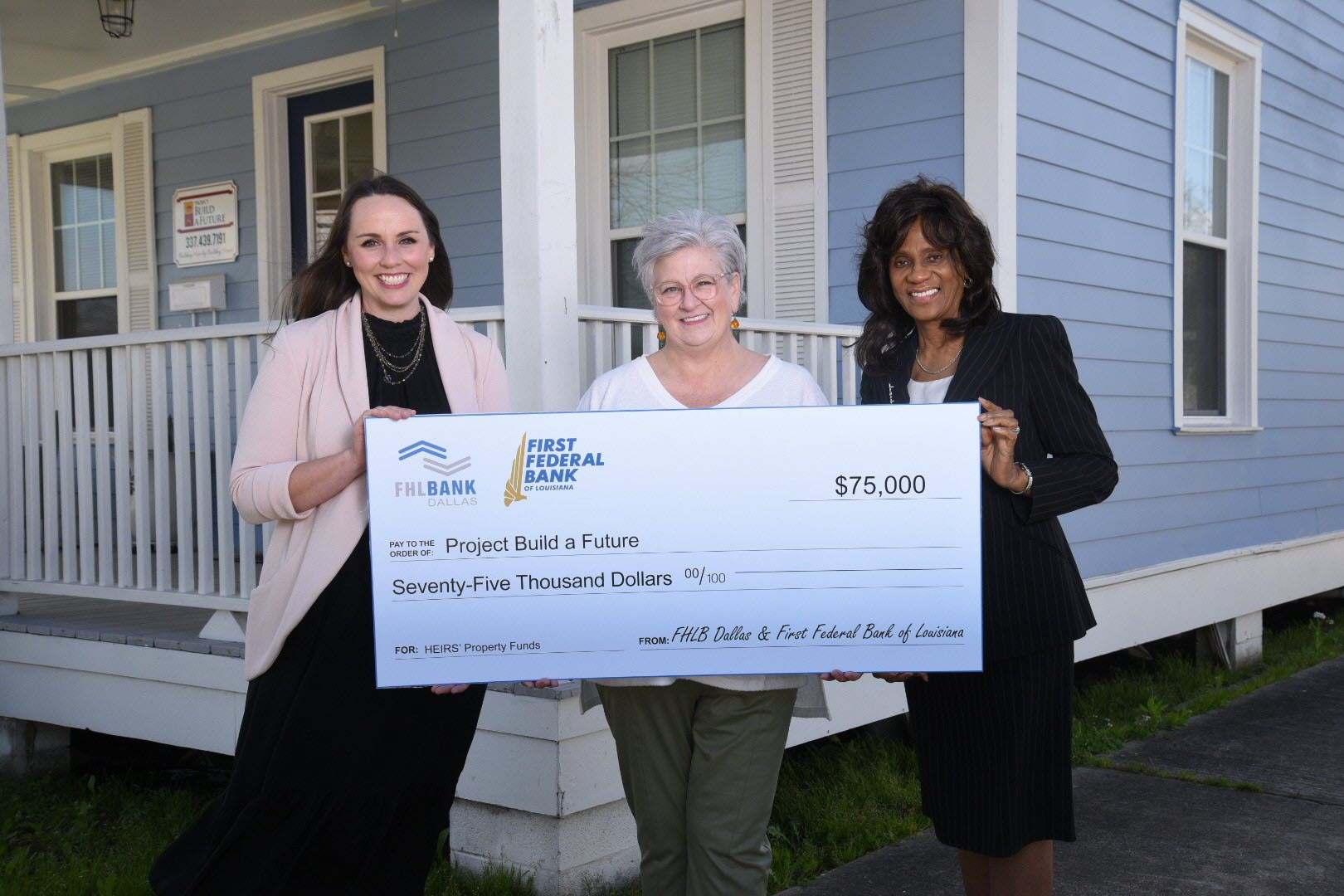 three women standing in front of a home holding a large check